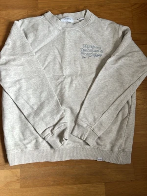 Les deux tröja - Ljusgrå sweatshirt från les deux med tryck fram och stort blått tryck på ryggen med texten 'Harajuku Teahouse & Boardshop'. Tröjan har rund hals, ribbade muddar och är tillverkad i mjuk bomullsblandning. Perfekt för en chill och avslappnad stil.