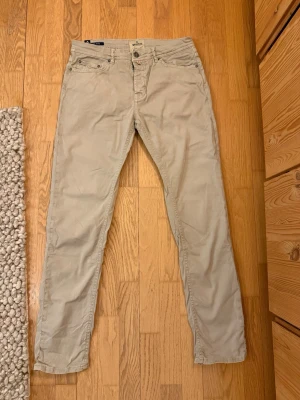 Beige jeans från Morris - Stilrena beige jeans från Morris med klassisk femficksdesign och raka ben, i en slim modell. Byxorna har snygga detaljer som logga på myntfickan och knapp, samt en liten orange söm vid gylfen. Strlek waist 32, längd 32. 