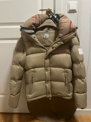 Beige dunjacka från Burberry - Snygg beige dunjacka från Burberry med klassiskt rutigt foder i huvan och dragkedja framtill. Jackan har två fickor med dragkedja och en patch med Burberry-logga på ärmen. Perfekt för kalla dagar och riktigt trendig look.