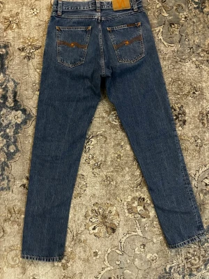 Nudie Jeans - Klassiska blå jeans från Nudie Jeans med raka ben och snygga kontrastsömmar. Jeansen har fem fickor och Nudie Jeans ikoniska broderi på bakfickorna. Perfekta för en avslappnad och stilren look.