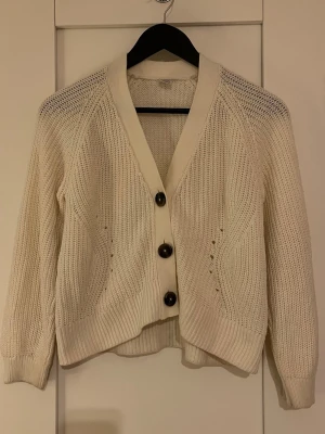 Vit stickad kofta med knappar - Säljer en vit stickad kofta med grov struktur och stora svarta knappar framtill. Koftan har långärmade ärmar och ribbade muddar vid ärmslut och nederkant. Perfekt att slänga över en t-shirt för en chill look.