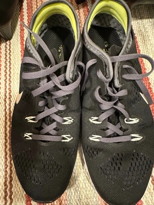 Nike Free 5.0 svarta sneakers - Nike Free 5.0 sneakers i svart mesh med grå detaljer och vit sula. Skorna har snörning och ventilerande ovandel med perforerat tyg. Insidan är ljusgul och sulan är flexibel för skön känsla vid träning. Swoosh-logga på sidan och klassisk rund tå.