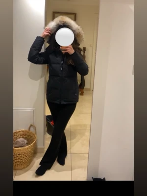 Svart Canada Goose dunjacka med päls - Säljer en svart dunjacka från Canada Goose med fluffig päls på huvan. Jackan har dragkedja framtill, två stora fickor och klassisk röd logga på ärmen. Perfekt för kalla dagar och har en snygg, normal passform. Den är använd max (3 gånger under en vinter). Köptes för 20 000kr i Stockholm. Jag är xs/s och den passar perfekt. 