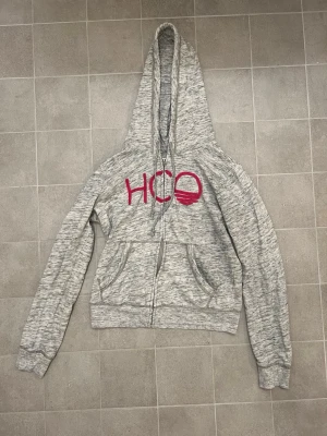 Hollister zip-up hoodie - Gråmelerad zip-up hoodie från Hollister i storlek S. Tröjan har dragkedja, huva med snörning och två fickor framtill. På bröstet finns en stor rosa HCO-logga i tyg. Perfekt för chill dagar och enkel att matcha med jeans eller mjukis.