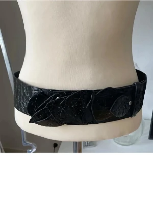 Svart bälte med bladdekor - svart bälte glansigt  med krokodilpräglad yta och dekorativa rörliga blad framtill.  bred form och cool struktur som ger en edgy vibe. Perfekt för dig som vill sticka ut med detaljer och mönster. Snygg att styla det mesta 