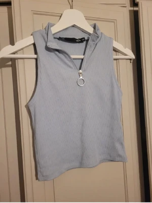 Ljusblå ribbad polotopp med dragkedja - Säljer en ljusblå ärmlös polotopp från 157 med ribbad struktur och en snygg silverfärgad dragkedja framtill med rund ringdetalj. Toppen har en hög krage och är gjord i mjukt syntetmaterial, perfekt för att styla med jeans eller kjol. Knappt använd. Tycker själv att den är mer som en small. 