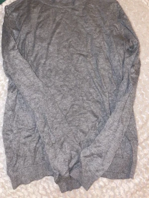 Grå stickad tröja från H&M Basics - Säljer en enkel och stilren grå stickad tröja från H&M Basics i storlek M. Tröjan har rund hals och långa ärmar med ribbade muddar vid ärmslut och nederkant. Perfekt för lager-på-lager under kyliga dagar.