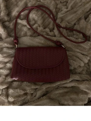 Vinröd axelväska med flätad design - Säljer en vinröd axelväska med lång rem och snygg flätad yta. Väskan har ett lock och är tillverkad i skinn, vilket ger en lyxig känsla. Perfekt storlek för det viktigaste och passar dig som gillar stilrena detaljer.