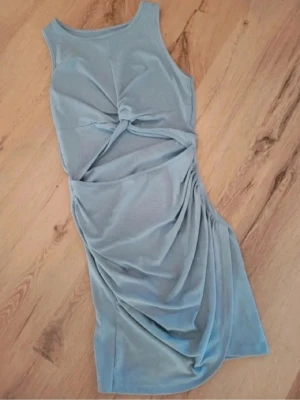 Ljusblå omlott kortklänning - Snygg ljusblå kortklänning med omlott-detalj och knyt framtill. Klänningen är ärmlös och har en cool draperad effekt längs midjan och höften. Materialet känns mjukt och stretchigt, troligen bomull eller polyester. Perfekt för dig som gillar trendiga och unika plagg. Aldrig använd. 