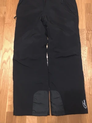 Svarta skidbyxor från Five-seasons - Svarta skidbyxor från Five-seasons med förstärkta partier vid bensluten och praktiska fickor med dragkedja. Byxorna har en rak passform och är gjorda i slitstarkt syntetmaterial, perfekta för kalla dagar och vinteraktiviteter, såsom att åka skidor eller snowboard. Snyggt broderad logga på benet.