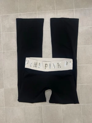 Svarta yoga pants från PINK Victoria's Secret - Svarta yoga pants från PINK Victoria's Secret med bred vit midjeresår dekorerad med silvriga paljetter som stavar 'PINK'. Byxorna har bootcut-modell och är gjorda i mjuk bomullsblandning med elastan för extra stretch. Perfekta för chill eller träning. Storlek M