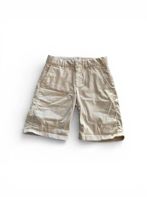 Beige shorts från Scotch & Soda - Snygga beige shorts från Scotch & Soda med klassisk passform och midwaist. De har fickor fram och bak, bälteshällor och stängs med knapp och dragkedja. Materialet är mjuk bomull som känns chill och passar perfekt till varma sommar dagar. Måtten är Längd 40cm  Bredd 34cm.