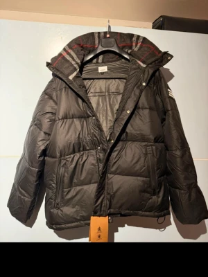 Svart pufferjacka Burberry med avtagbara ärmar - Svart pufferjacka från avtagbara ärmar så du kan använda den som väst. Jackan har huva med rutigt foder, dragkedja och knappar framtill samt två sidofickor med dragkedja. Perfekt för kalla dagar och riktigt snygg stil. Stor Burberry logga på armen.