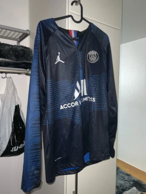 PSG x Jordan långärmad träningströja - Säljer en mörkblå långärmad träningströja från PSG och Jordan med halv dragkedja. Tröjan har blå och svarta ränder, PSG-logga på bröstet och Jordan-logga i vitt. Materialet är syntet och perfekt för fotbollsträning.