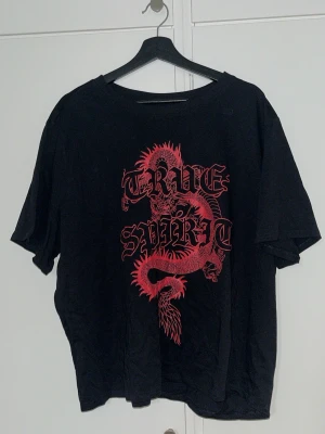 Svart t-shirt med röd drake - Cool svart t-shirt med ett stort rött draktryck och texten 'TRUE SPIRIT' på framsidan. T-shirten har rund hals och korta ärmar, perfekt för dig som gillar streetstyle och grafiska prints. Materialet känns mjukt och skönt mot huden. Köpt från H&M och är aldrig använd