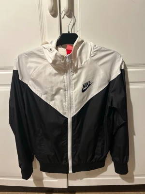 Nike svartvit vindjacka med huva - Nike vindjacka i storlek M men passar även en S. Jackan har huva, hel dragkedja och två sidofickor. Lätt och sportig. Materialet är tunt och passar perfekt under en vanlig höstdag. 