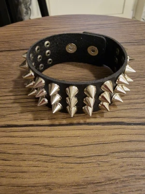 Svart nitad choker med spikes - Säljer en svart choker med stora silvriga spikes och metallnitar runt hela bandet. Chokern är tillverkad i PU skinnimitation och har tryckknapp för enkel stängning. Perfekt för dig som vill ha en edgy och alternativ stil. 23 cm lång. Bredd 3 cm.