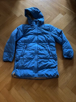 Blå vinterjacka från Tretorn med huva - Blå vinterjacka från Tretorn med huva och dragkedja framtill. Jackan har två sidofickor med dragkedja och är quiltad för extra värme. Insidan är beige och jackan har lång ärm med elastiska muddar. Perfekt för kalla dagar.