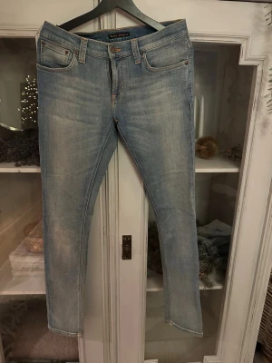Ljusblå jeans från Nudie Jeans. Modell long John  - Säljer ett par ljusblå skinny jeans från Nudie Jeans med klassisk femficksdesign och snygga slitningar. Jeansen har orange kontrastsömmar, läderpatch bak och är tillverkade i mjukt bomullsdenim. Perfekta för dig som gillar en smal passform och stilren look.