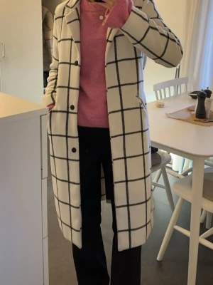 Vit rutig kappa med knappar - Säljer en snygg vit kappa med svart rutigt mönster och klassisk krage. Kappan har knappar framtill och två fickor på sidorna. Perfekt för dig som gillar stilrena och trendiga ytterplagg med lite längre passform.