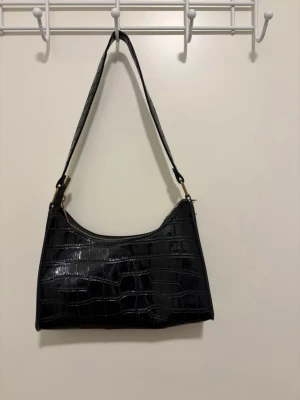 Svart axelväska i croco-mönster - Snygg svart axelväska med croco-mönstrad yta och glansig finish. Väskan har en rak, avlång form och ett brett axelband med metallfästen. Stängs med dragkedja upptill och är perfekt för dig som gillar stilrena detaljer.