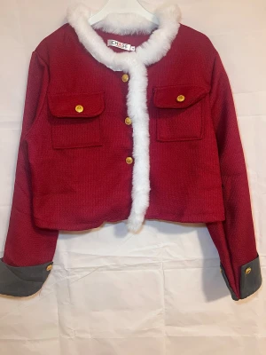 VinRöd jacka med fuskpäls från Chase - Säljer en röd jacka från Chase med vit fuskpäls runt kragen och längs knäppningen. Jackan har guldknappar, två stora bröstfickor och kontrasterande gråa muddar vid ärmsluten. Croppad modell som ger en cool vibe.