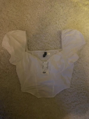 Vit blus med puffärmar från H&M - Supersöt vit blus från H&M med puffiga korta ärmar och fyrkantig urringning. Toppen har smock baktill för extra komfort och en figurnära passform. Perfekt att styla med jeans eller kjol för en trendig look.