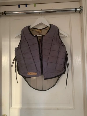 Grå säkerhetsväst från Racesafe XL barnstorlek - Säljer en grå säkerhetsväst från Racesafe i storlek XL, barnstorlek. Västen har quiltad design, dragkedja framtill och snörning i sidorna för justerbar passform. Bra skick endast fläckig på många ställen, kan säkert gå bort vid tvätt. Märkt med Racesafe-logga på framsidan.