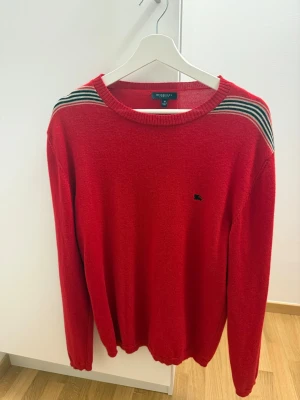 Röd stickad tröja från Burberry - Säljer en röd stickad tröja från Burberry med klassiska ränder i svart, vit och grön på axlarna. Tröjan har rund hals, lång ärm och en liten broderad logga på bröstet. Perfekt för dig som gillar stilrena detaljer och snygg design.