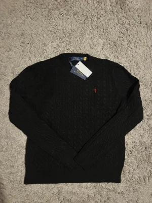 Svart stickad tröja Polo Ralph Lauren - Svart kabelstickad tröja från Polo Ralph Lauren med klassisk röd logobroderi på bröstet. Rund hals och långa ärmar, tillverkad i mjuk bomull. Perfekt för dig som gillar stilrena och tidlösa plagg med en sportig touch.