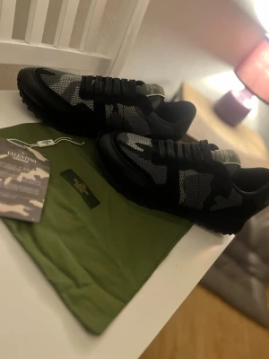 Valentino Rockrunner sneakers svart/grå - Snygga Valentino Rockrunner sneakers i svart och grått med camouflagemönster. Skorna har snörning, rund tå och platt sula. Materialmix av skinn och textil ger en cool och modern vibe. Perfekt för dig som gillar exklusiva märken och streetstyle.