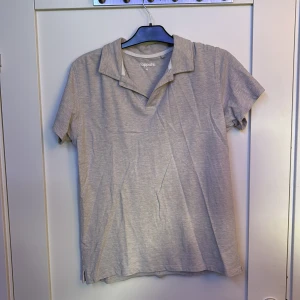 Grå piké t-shirt från Kappahl - En stilren beige piké t-shirt från Kappahl. Pikén har klassisk krage och korta ärmar, perfekt för en clean och avslappnad look. Passformen är normal. Färgen syns bäst på sista bilden