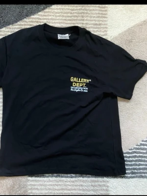 Svart t-shirt Gallery Dept med biltryck - Svart t-shirt från Gallery Dept. i bomull med coolt tryck på ryggen som visar en röd sportbil och palmer. Framsidan har en liten gul och vit logga på bröstet. Klassisk rund hals och korta ärmar, perfekt för dig som gillar streetwear och grafik.