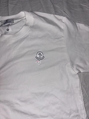 Vit Moncler t-shirt med logga - Snygg vit t-shirt från Moncler i bomull, med klassisk rund hals och korta ärmar. På bröstet sitter Monclers ikoniska logotyp i vitt och svart. Enkel och stilren design som funkar till allt. Perfekt för dig som gillar exklusiva märken och clean look.