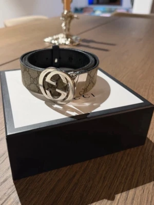 Gucci beige GG-mönstrad bälte - Säljer ett snyggt Gucci-bälte med klassiskt GG-mönster i beige och grå toner. Bältet har en stor, rund silvrig spänne med Gucci-logga och svarta detaljer på insidan. Materialet är slitstark canvas med läderdetaljer. Perfekt för att lyfta din outfit.