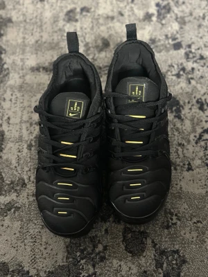 Nike Air Vapormax Plus Svart/Guld - Nike Air Vapormax Plus sneakers i svart med detaljer i guld. Skorna har en chunky, genomskinlig sula med synliga luftkuddar, snörning och en syntetisk ovandel med vågiga mönster. Snygga guldinslag på sidan och tungan ger extra edge till looken.