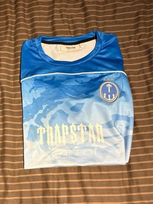 Blå camo t-shirt från Trapstar - Säljer en blå t-shirt från Trapstar med coolt camouflagemönster och tryck både fram och bak. På ryggen står det 'Trapstar' och siffran 23 i stor stil. Framsidan har logga med tre stjärnor och texten 'It's a secret'. Perfekt för dig som gillar streetwear. S men passar M