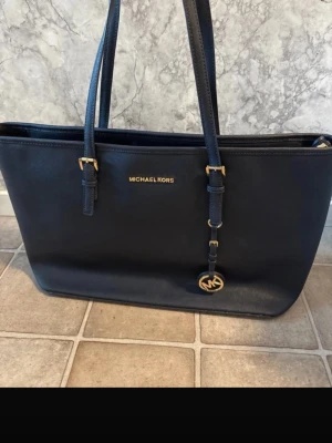 Michael Kors Mörkblå axelväska  - Snygg mörkblå axelväska från Michael Kors i skinn med guldfärgade detaljer och logotyp framtill. Väskan har två långa axelremmar och praktiskt innerfoder med flera fack och dragkedja. Perfekt för dig som vill ha både stil och funktion.