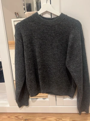 Mörkgrå stickad tröja med rund hals - Mysig mörkgrå stickad tröja med rund hals och långa ärmar. Tröjan har en enkel och klassisk design utan mönster eller detaljer, perfekt för att hålla dig varm under kyliga dagar. Passar dig som gillar en chill och stilren look.