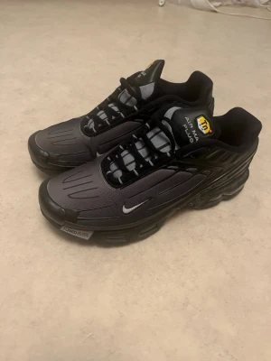 Nike Air Max Plus svarta sneakers - Nike Air Max Plus sneakers i svart och grått med Tuned Air-dämpning. Snyggt futuristiskt mönster på ovandelen, små silvriga swoosh-loggor och svarta snören. Platt sula och rund tå, tillverkade i syntetmaterial. Tn-logga på plösen och bak på hälen.