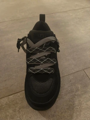 Svarta UGG sneakers med grå snören - Snygga svarta sneakers från UGG med coola grå och svarta snören med mönster. Ovandelen är i mesh och mocka för extra stil och komfort. Skon har rund tå och platt sula, perfekt för dig som gillar streetwear och vill ha något som sticker ut.