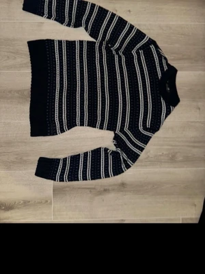 Svartvit randig stickad tröja - Säljer en svart stickad tröja med vita ränder och diskret mönster. Tröjan har lång ärm, rund hals och breda muddar vid ärmslut och nederkant. Perfekt för dig som gillar klassisk stil med en twist. Passar dig som vill ha något mysigt och snyggt till kyliga dagar.