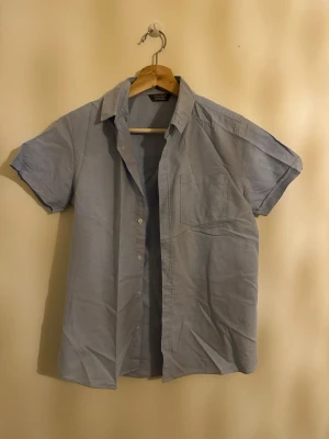 Ljusblå kortärmad skjorta Primark - Säljer en ljusblå skjorta från Primark med korta ärmar och klassisk krage. Skjortan har knappar framtill och en bröstficka. Perfekt för dig som gillar enkel och stilren look. Materialet är mjukt och lätt bomull.