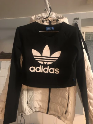 Svart croppad långärmad topp Adidas - Säljer en svart croppad långärmad topp från Adidas med stort vitt tryck av Adidas logga på bröstet. Toppen har rund hals och är tillverkad i mjuk bomull, perfekt för en sportig och chill stil.