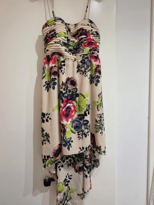Blommig långklänning från Vila - Säljer en superfin långklänning från Vila med blommigt mönster i rött, grönt, blått och svart på beige botten. Klänningen har smala axelband, omlottliknande överdel och faller snyggt med asymmetrisk nederkant. Perfekt för sommarens alla tillfällen!