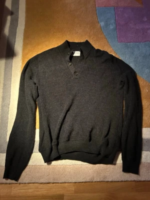 Mörkgrå stickad tröja Bruun & Stengade - Snygg mörkgrå stickad tröja från Bruun & Stengade i storlek L. Tröjan har en krage med två knappar vid halsen och ribbade muddar vid ärmslut och nederkant. Perfekt för dig som gillar stilrena och klassiska plagg. Vid midjan är den sydd kortare än originalet (var överdrivet lång tidigare) 