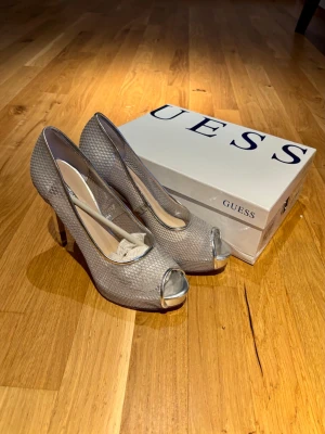 Silvriga pumps från Guess med klack - Säljer ett par silvriga pumps från Guess med snyggt glittrigt mönster och metalliska detaljer vid öppna tån. Skorna har hög klack och rundad form, perfekt för dig som gillar att sticka ut. Materialet är syntet med en skimrande finish och insida i beige ton. Endast provade