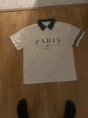 Vit polotopp med Paris-tryck - Säljer en vit polotopp med mörkgrön krage och trycket 'PARIS FRANCE' på framsidan. Toppen har korta ärmar med två mörkgröna ränder på ena ärmen. Materialet känns som polyester och toppen har en klassisk piké-look.