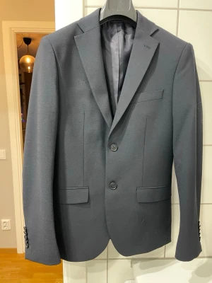 Mörkgrå kostym från Pour Homme - Säljer sonens mörkblå kostym från Pour Homme med klassisk tvåknäppt kavaj och matchande byxor. Kavajen har smala slag, innerfoder i blankt tyg och raka fickor med lock. Perfekt för dig som vill ha en stilren och tidlös look. Han har bara använt dem vid ett tillfälle (vid skolavslutningen i 9:an i somras) så den är typ oanvänd. Fickorna på kavajen är tex inte uppsprättade. Min son var runt 165 - 170cm när han hade den. Skick som ny.