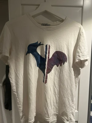 Vit Moncler t-shirt - Vit t-shirt från Moncler med ett stort grafiskt tryck av en tupp i blått och rött på framsidan. Klassisk rund halsringning och korta ärmar. Moncler-logga på vänster ärm. Tillverkad i mjuk bomull för en skön känsla. Tröjan har krympt i tvätten va L men sitter mer som S nu